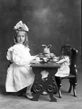 A girl with a bow in her hair, having tea with her toy rabbit and doll